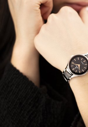 Persona que lleva un reloj de pulsera analógico plateado con esfera negra y marcadores de hora dorados, manos cruzadas cerca de su rostro, vistiendo un suéter negro.