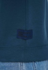 Mørkeblå hettegenser nederst med et sydd merke som står "DEDICATED TO DENIM PERFECTION" over lys blå jeans med belte.