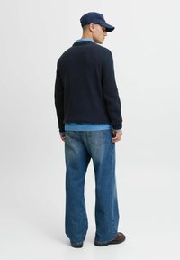 Cardigan texturé marine sur chemise bleu clair, associé à un pantalon en denim bleu à jambes larges et des chaussures noires, vu de dos.