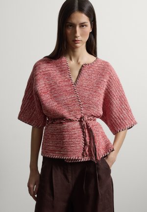 Vrouw in een roze gebreide top met brede mouwen en een gevlochten riem, gecombineerd met donkerbruine, hooggetailleerde broek, staand tegen een effen achtergrond.