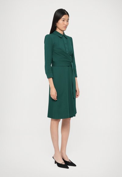 Πράσινο φόρεμα τύπου wrap από μαλακό ύφασμα, με γιακά, μακριά μανίκια, κουμπιά εμπρός και ζώνη στη μέση. Φοριέται με μαύρες γόβες.