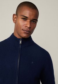 Mornarska zip-up veste s visokim ovratnikom, izrađena od mekog materijala. Sadrži mali logo na prsima i glatku, teksturiranu završnu obradu.