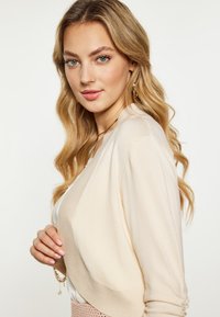 Beige leichter Cardigan mit weicher Textur, offener Front und gerafften Ärmeln, kombiniert mit einem weißen Oberteil und einem pinkfarbenen strukturierten Rock.