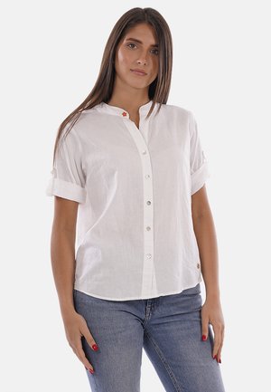 Femme aux longs cheveux bruns portant une chemise blanche boutonnée avec les manches retroussées et un jean bleu, debout devant un fond uni.