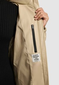 Béžová puffrová bunda s prešívanou textúrou a čiernou zipsovou vreckom. Štítok vo vnútri hovorí: "Teplé objatie vyrobené z látky." Viditeľný čierny rebrovaný top.