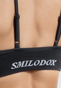 Zwarte sportbeha met een ribbelstructuur, voorzien van verstelbare banden en witte geborduurde "SMILODOX" branding op de band.