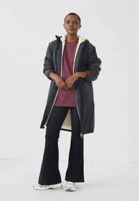 Abrigo largo negro con forro blanco de peluche, combinado con una camisa granate y pantalones acampanados negros. Zapatillas plateadas complementan el conjunto.