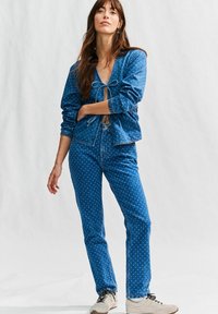 Denim kombinacija s prilagođenim sakoom s točkicama koji ima vezanje sprijeda i elastične manžete, uparen s odgovarajućim trapericama s točkicama. Svijetloplava boja.