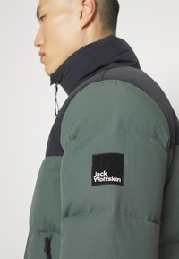 Grøn og sort pufferjakke med høj krave, der har en firkantet sort lap med hvid "Jack Wolfskin" logo på venstre skulder.