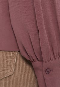 Bluza dugih rukava u mauve boji, s nabranim rukavima u obliku puff i kopčanjem na manžetama, u kombinaciji s teksturiranim bež kordom.