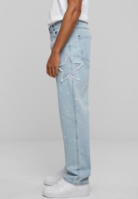 Light blue ripped denim jeans featuring a frayed star design on the thigh and white paint splatter accents, styled with white sneakers.