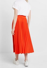 Falda midi con pliegues en un brillante color naranja, hecha de una tela ligera. Presenta una cintura alta y cae por debajo de la pantorrilla. Se lleva con una camisa blanca y zapatillas.