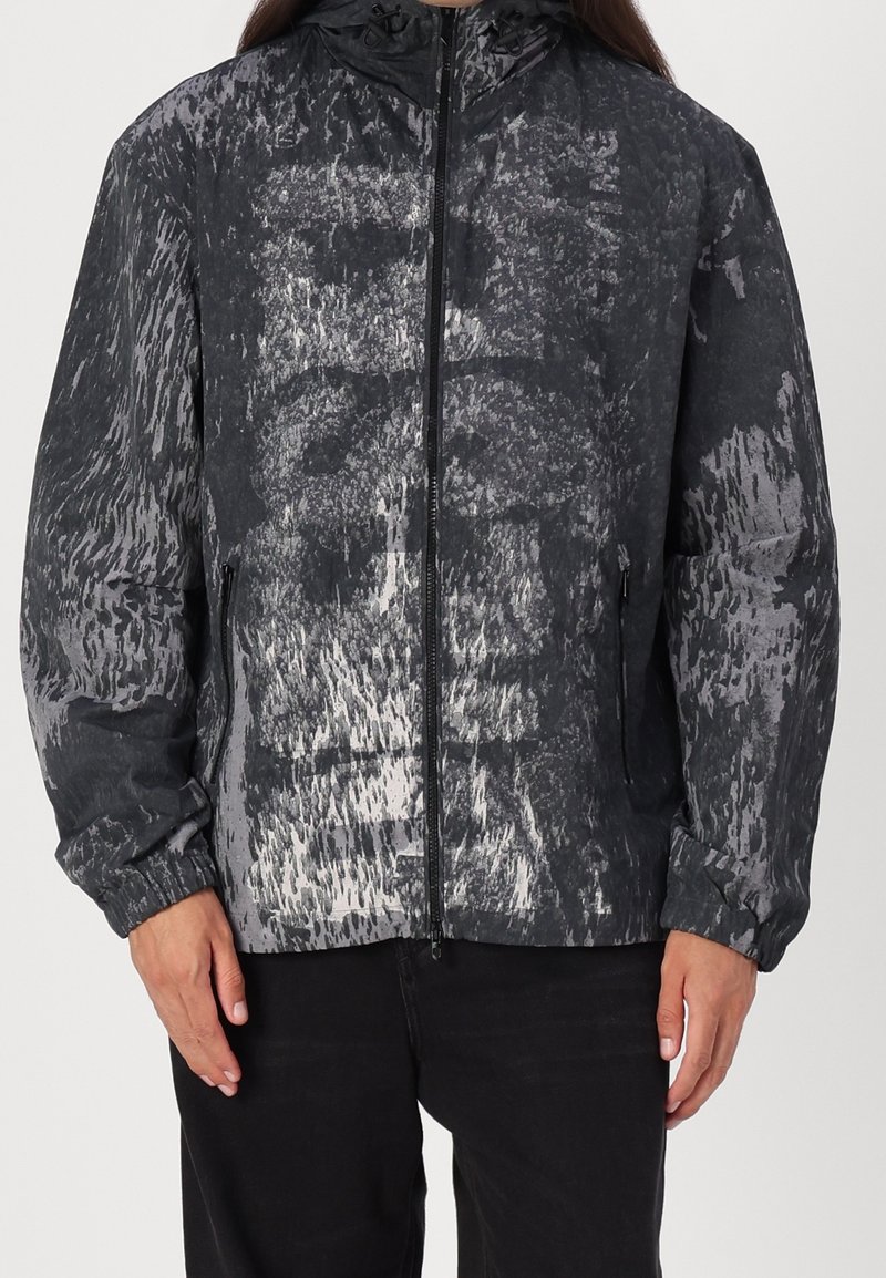 Personne portant une veste à capuche noire et grise à motif abstrait, avec fermeture éclair frontale et poches latérales zippées.