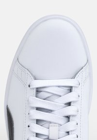 Gros plan sur la pointe d'une basket blanche avec des lacets blancs noués en motif croisé et des coutures visibles sur les côtés.