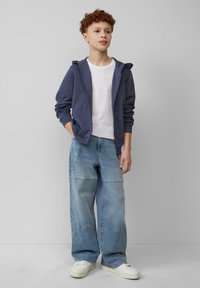 Marineblauwe ritscrewneck hoodie met kangoeroezakken over een wit T-shirt; gecombineerd met losse lichtblauwe jeans met stiknaaddetails en witte sneakers.