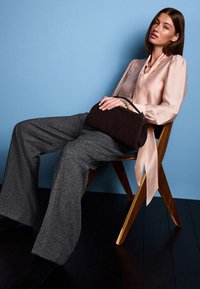 Brūna zamšādas rokassoma ar izliekta formu, valkāta kopā ar gaiši rozā zīdainu blūzi un pelēkām teksturētām platām biksēm, sēžot uz koka krēsla.