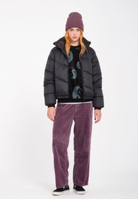 Giacca a vento nera con sezioni a trapunta orizzontale, indossata sopra un maglione nero a fantasia e pantaloni di velluto a coste viola, completata da un berretto viola.