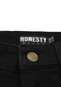 Sorte denim jeans med en messingknap og et mærke, der står "ÆRLIGHED REGNER." Stoffet har en tekstur med synlige sømme.