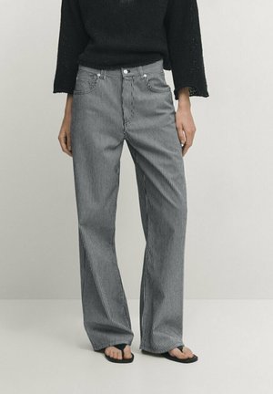 Kobieta ubrana w czarno-białe spodnie w szerokie prążki z wysokim stanem, czarny sweter dzianinowy i czarne klapki stojąca na tle jednolitego tła.