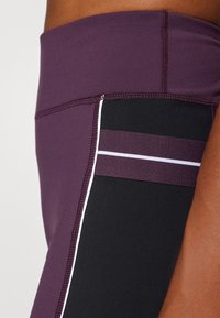 Sportos leggings magas derékkal, lila anyag fekete oldalpanelekkel, fehér csíkokkal és kontrasztos varrással kiemelve.