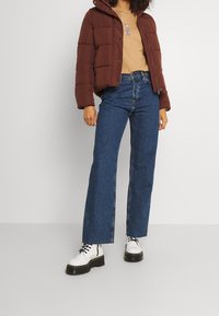 Brun puffjacka, beige grafisk t-shirt, blå utsvängda jeans och vita plattformsstövlar med svarta sulor. Texturerade material och lagerdesign.