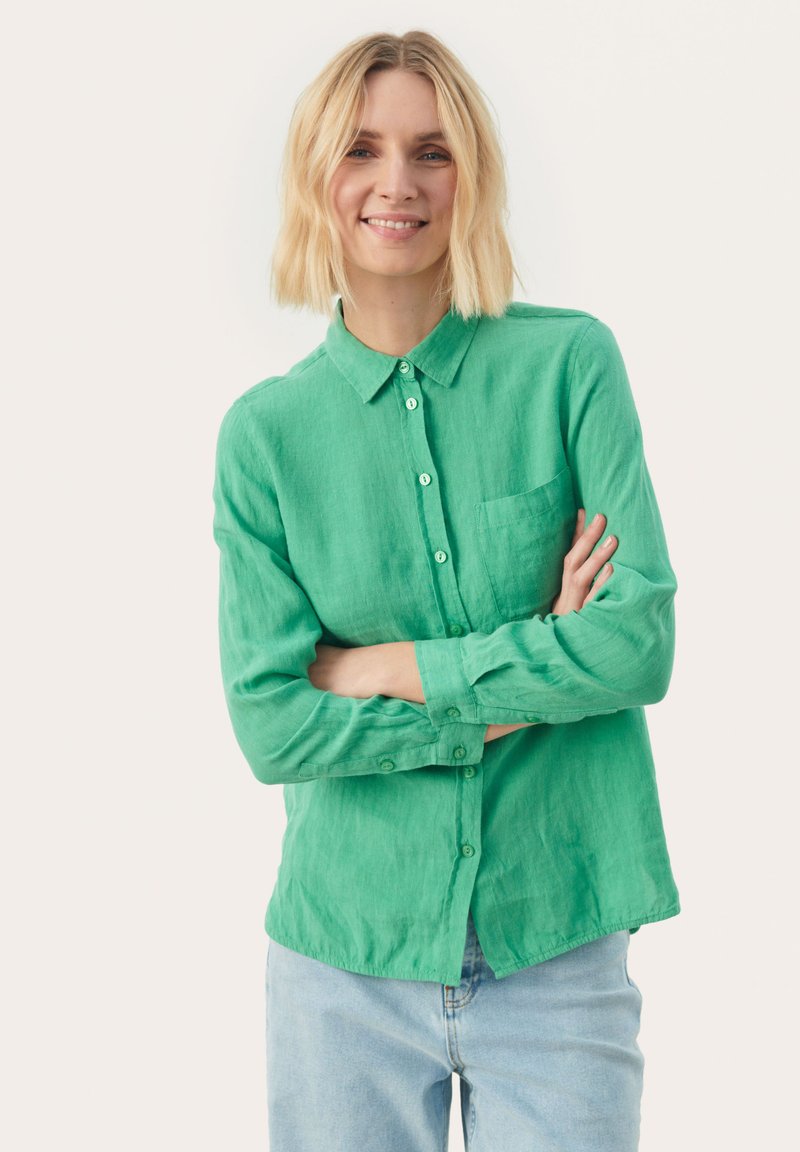 Grøn skjorte med knaplukning og krave, lange ærmer, frontlomme og et tekstureret stof. Kombinationen af skjorten er med lyseblå jeans.