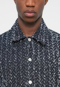 Veste texturée bleu marine avec des mouchetures blanches, fermeture à boutons sur le devant et col pointu. Caractérisée par un tissu tissé à motifs et deux boutons.