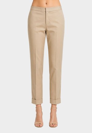 Pantaloni beige su misura alla caviglia con orli risvoltati indossati con tacchi beige aperti con cinturino alla caviglia su uno sfondo neutro.