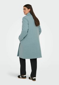 Gai zils vilnas mētelis ar augstu apkakli, ar garām piedurknēm un sānu kabatām, valkāts virs melniem biksēm un sporta apaviem.