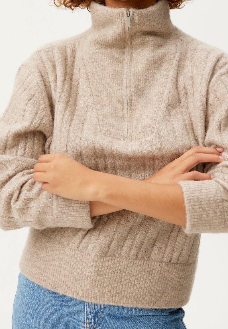 Personne portant un pull beige en maille côtelée avec une demi-fermeture éclair et des manches longues, les bras croisés, assorti à un jean en denim bleu.
