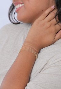 Pulsera de cadena de oro con múltiples hilos delicados, llevada en una muñeca. Textura suave, diseño elegante, que complementa una camisa gris.