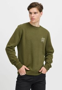 Sudadera verde oliva, con cuello redondo, puños y dobladillo acanalados, que presenta una impresión gráfica blanca de una pancarta en la parte superior izquierda. Material de mezcla de algodón.