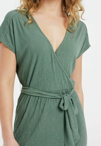 Vestido verde de tejido texturizado con escote en V profundo, mangas cortas y un cinturón atado en la cintura, con un patrón de rayas verticales.