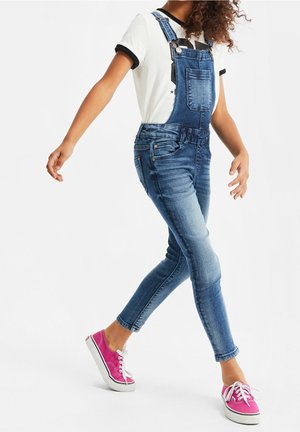 Lapsi kävelee pukeutuneena sinisiin farkkuhousuihin, valkoiseen t-paitaan mustalla reunuksella ja kirkkaan vaaleanpunaisiin tennareihin.