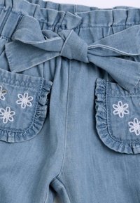 Shorts en denim de couleur bleu clair, avec une taille froncée, un nœud papillon et des poches à rabat froncées ornées de broderies florales blanches.