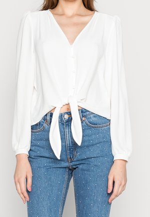 Vrouw draagt een witte, lang mouw blouse met knopen, gestrikt in de taille, gecombineerd met hoog getailleerde blauwe spijkerbroek met kleine witte stippen.