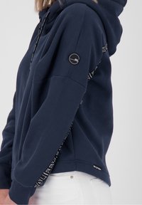Sudadera azul marino con una textura suave, con capucha ajustable y detalles decorativos a lo largo de las mangas. Incluye una insignia de la marca en el brazo.
