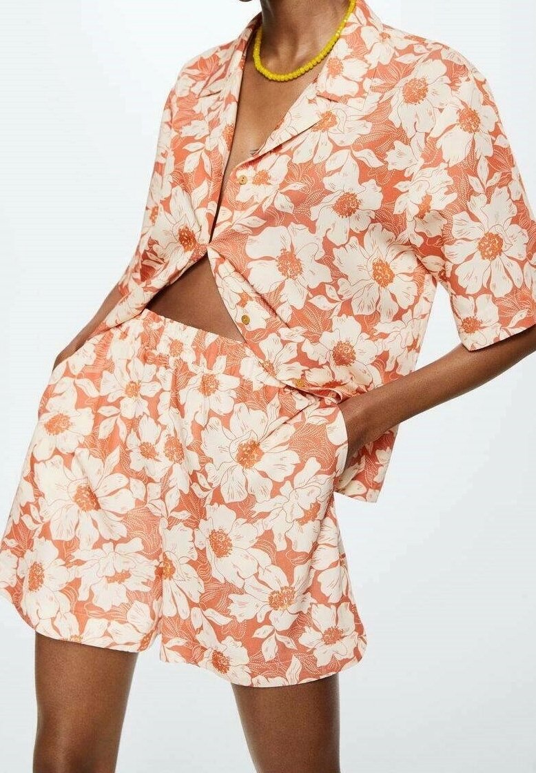 Chemise courte à manches courtes boutonnée orange et blanche à motifs floraux, avec un short assorti à taille élastique, portée avec un collier de perles jaunes.