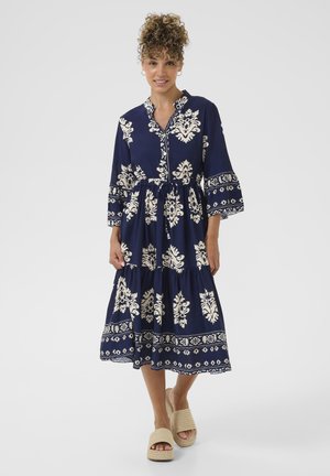 Abito blu navy a strati con motivi floreali bianchi, maniche lunghe, colletto e vita elastica. Indossato con sandali espadrilles beige.