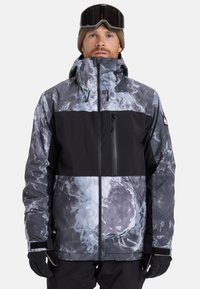 Mann trägt eine graue und schwarze gemusterte Winterjacke mit Kapuze, schwarze Handschuhe, eine braune Strickmütze und schwarze Skibrillen auf der Stirn.