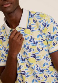 Korte mouwen polo met een patroon van gele, blauwe en groene bladeren en citroenen. Voorzien van een witte kraag met blauwe en gele accenten. Textuurstof.