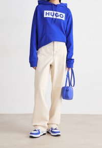 Modrá mikina s bílým logem "HUGO", béžové kalhoty s širokými nohavicemi, modrá crossbody taška a tenisky s modro-bílými akcenty. Hladká textilie.