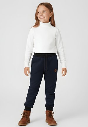 Młoda dziewczyna uśmiechająca się, ubrana w biały sweter z golfem, granatowe spodnie dresowe ze sznurkiem oraz brązowe wiązane buty, stojąca na jednolitym tle.