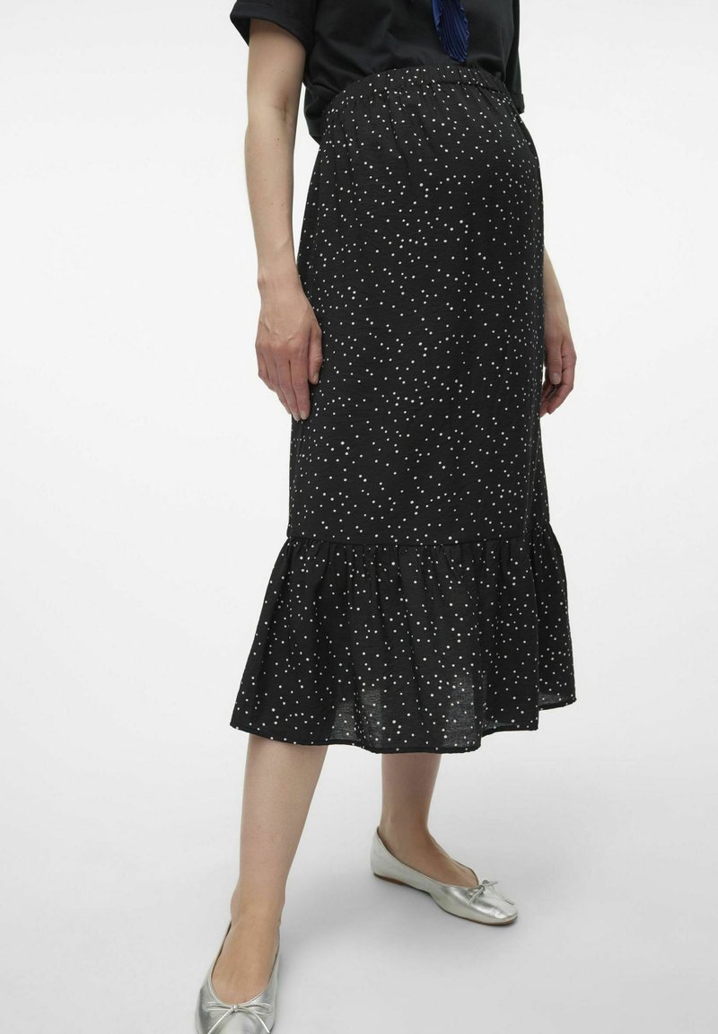 Jupe noire avec des pois blancs, longueur genou, bas évasé, taille élastique. Associée à des ballerines argentées pour un look complet.