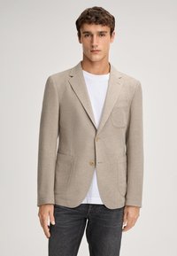 Blazer beige con tessuto texturizzato, design monopetto, revers a punta, due tasche frontali e bottoni marrone chiaro, indossato sopra una camicia bianca.
