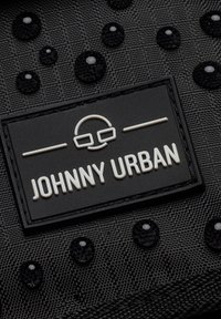 Czarna teksturowana tkanina z błyszczącymi czarnymi kroplami oraz białym wytłoczonym logo "JOHNNY URBAN" na prostokątnej czarnej metce.