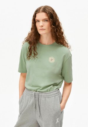 Femme aux longs cheveux bouclés portant un t-shirt vert clair avec un motif soleil et un pantalon de survêtement gris, debout les mains dans les poches sur fond blanc.