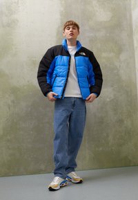 Blå og sort puffjakke med lynlås, udstyret med sidelommer. Båret over en hvid T-shirt med løse blå jeans og multicolore sneakers.