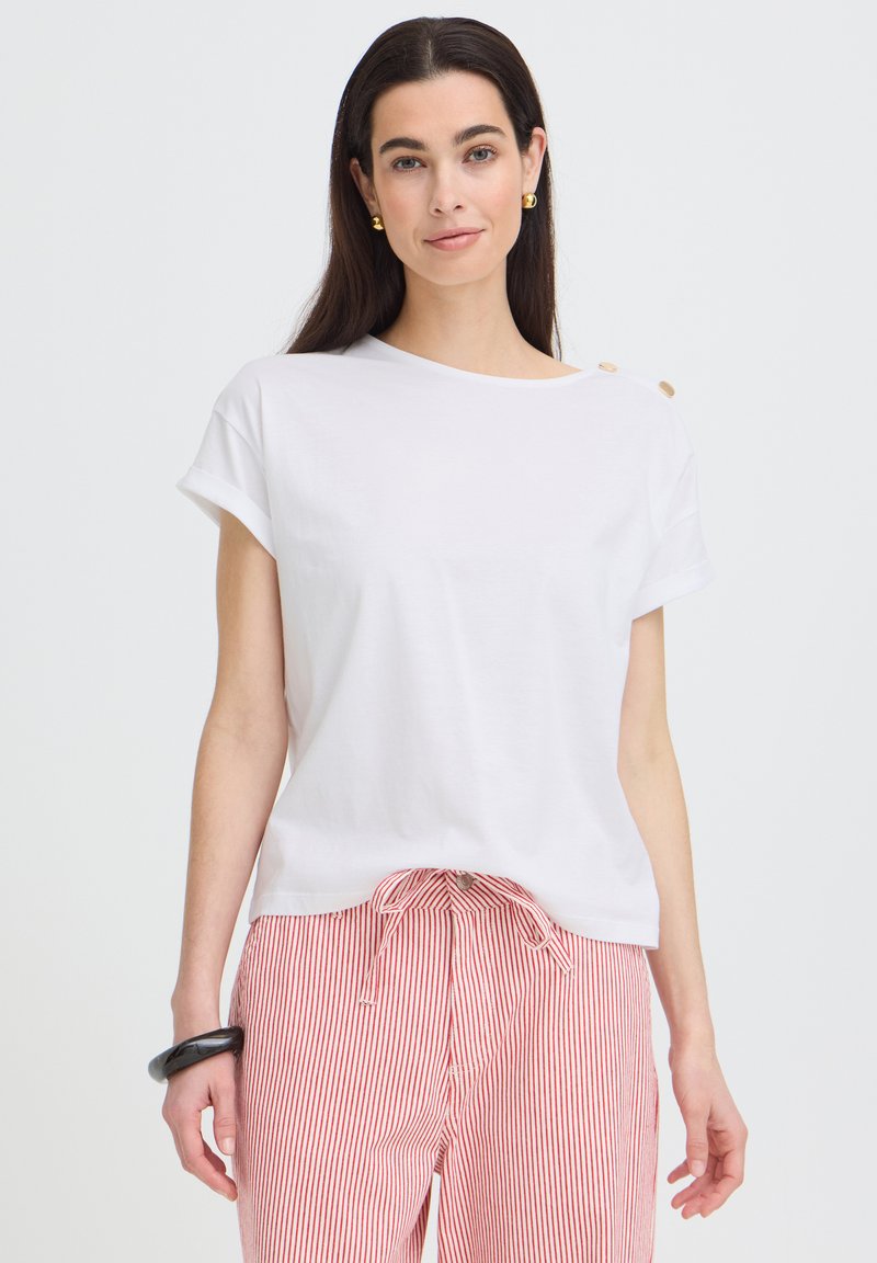 Femme aux longs cheveux foncés portant un haut blanc à manches courtes avec des boutons et un pantalon rayé rouge et blanc, accessoirisée de boucles d'oreilles en or et d'un bracelet noir.