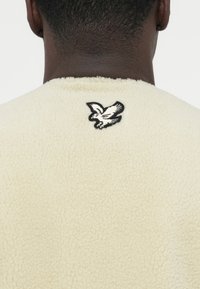 Felpa in pile color crema con una texture morbida, con un logo aquila ricamato in nero sul retro del collo.